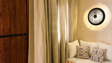 Una habitación con puerta de madera, cortinas color crema, un sofá blanco con cojines estampados, una pequeña mesa redonda con una cesta y vasos, y una lámpara fijada a la pared.