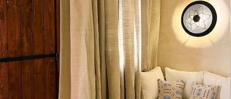 Una habitación con puerta de madera, cortinas color crema, un sofá blanco con cojines estampados, una pequeña mesa redonda con una cesta y vasos, y una lámpara fijada a la pared.
