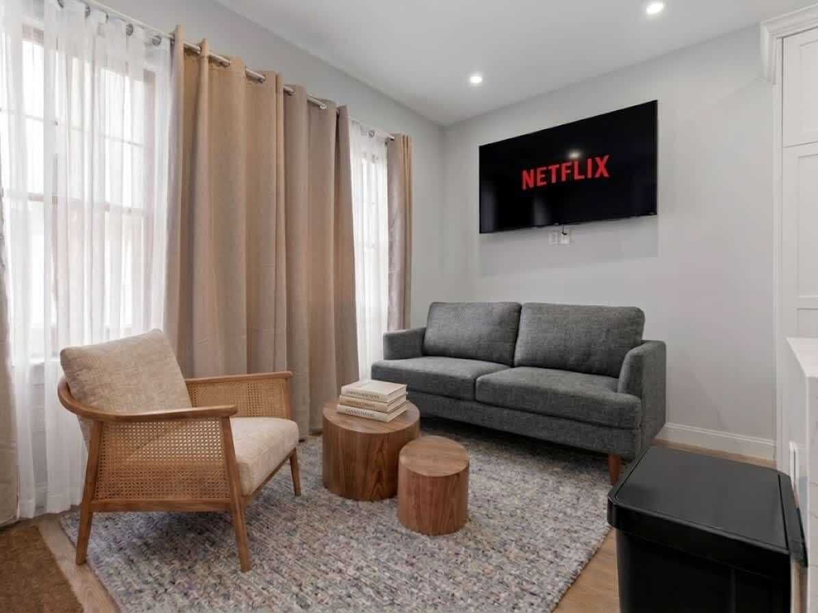 A living room with a grey sofa, a wicker chair, a small wooden table, and a TV mounted on the wall.