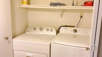 A laundry room with two white front-loading washers and dryers, a shelf above the dryers, and a door to the left.