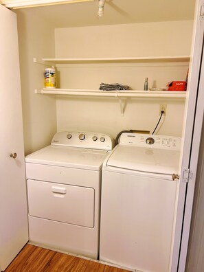 Une buanderie équipée de deux lave-linge et deux sèche-linge à chargement frontal, tous deux de couleur blanche, une étagère au-dessus des sèche-linge et une porte située à gauche.