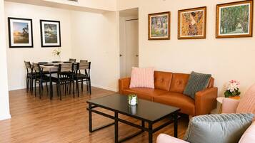 Un salon avec un canapé en cuir marron, une table basse et un coin repas comprenant une table et des chaises. Des tableaux encadrés ornent le mur.