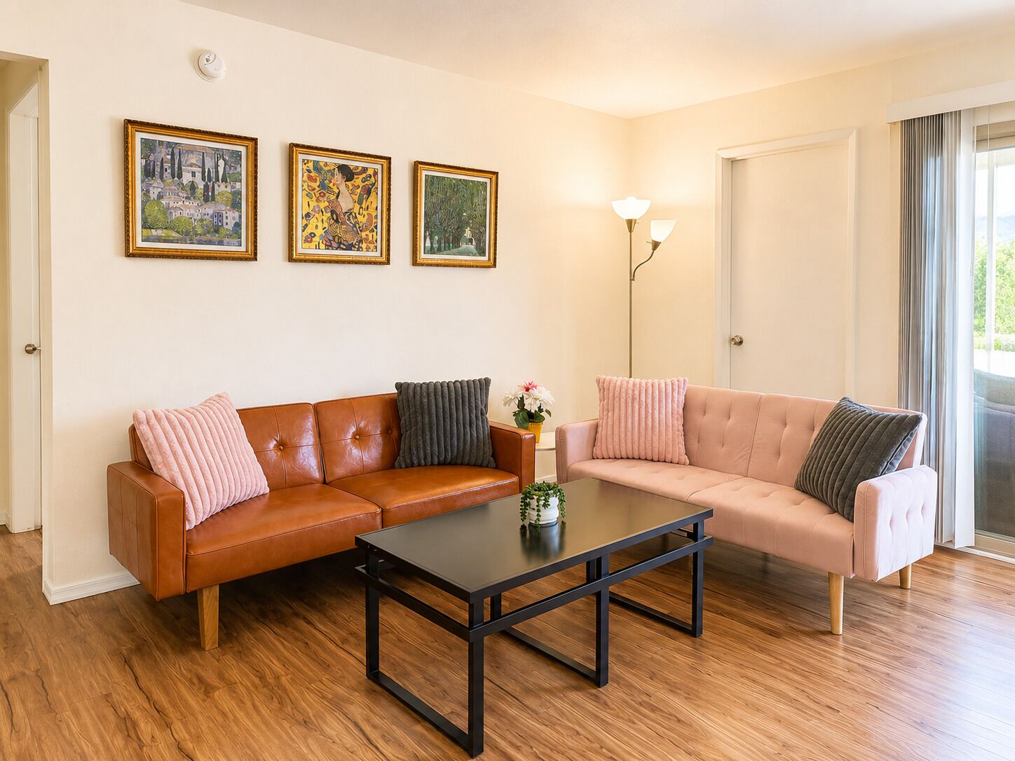 Un salon avec un canapé en cuir marron, un canapé rose pâle, une table basse noire et trois œuvres d’art encadrées accrochées au mur.