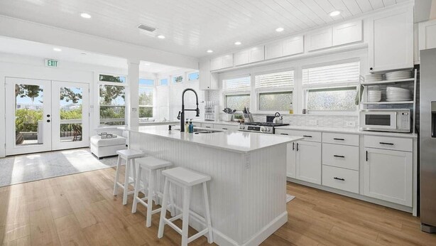 Una cocina moderna con armarios blancos, una isla central con taburetes, un refrigerador de acero inoxidable y vistas a través de puertas de cristal.