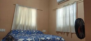 Een slaapkamer met een bed, twee ramen met gordijnen, een airconditioner en een ventilator aan de muur.