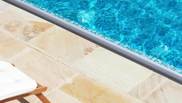 Uma área na beira da piscina com piso em azulejos e duas espreguiçadeiras de madeira com almofadas brancas.