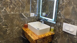 Un baño con pared de mármol, tocador de madera, lavamanos blanco y dos espejos.