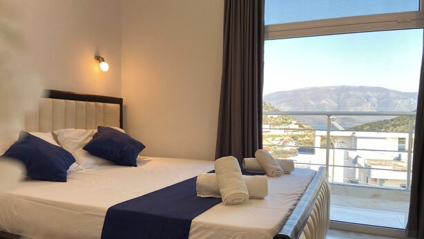 Habitación de hotel con cama, mesita de noche, ventana con vistas al mar y a las montañas, y balcón.