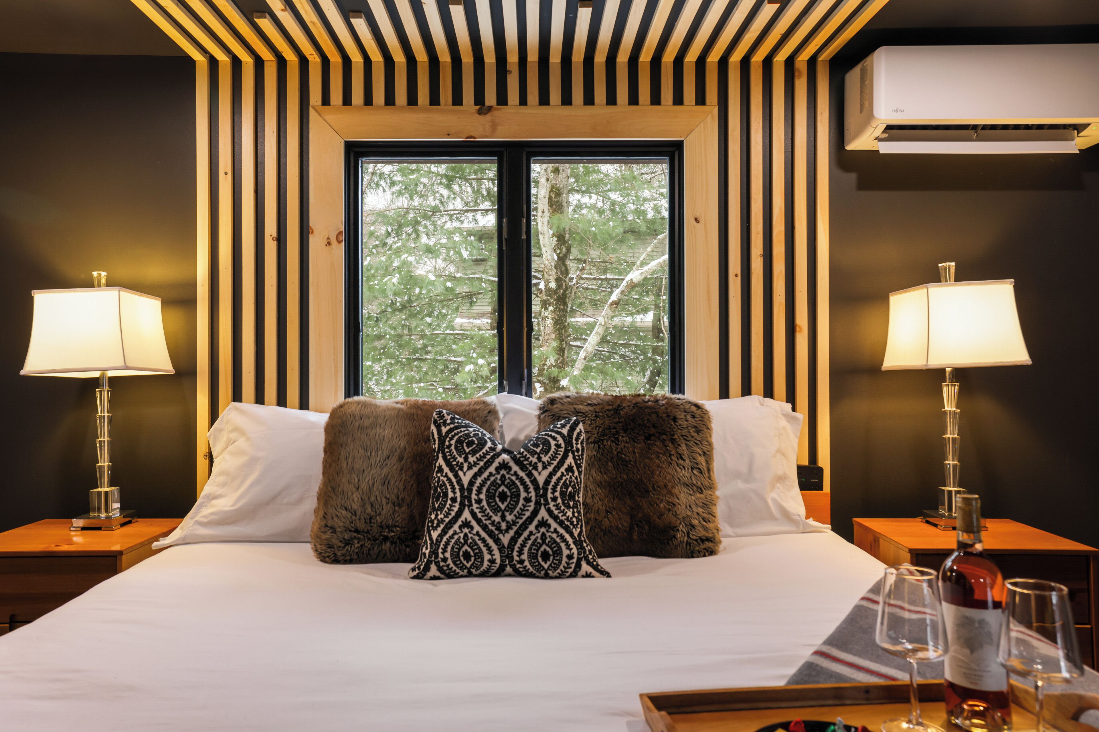 Habitación de hotel con una cama grande, dos lámparas de noche, un ventanal con vistas a árboles y un equipo de aire acondicionado en la pared.