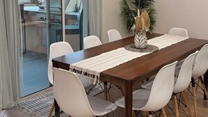 Un coin repas avec une table en bois, des chaises blanches, un centre de table orné de plumes de paon et une vue sur un barbecue extérieur, visible à travers des portes vitrées.