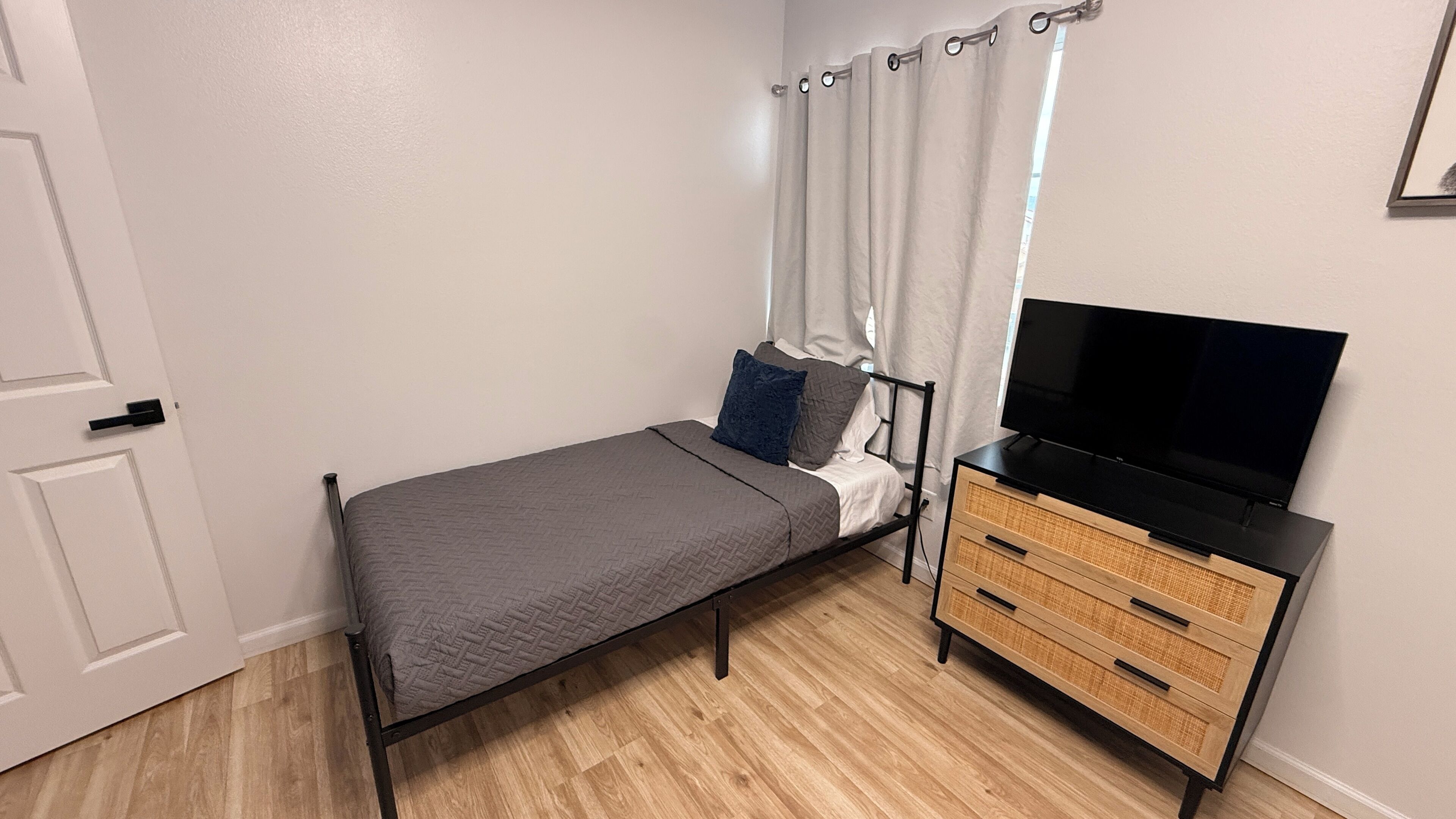 Une petite chambre à coucher, bien rangée, avec un lit simple, un téléviseur posé sur une commode en bois et une porte dotée d’une poignée.