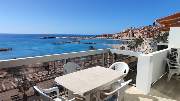 En balkon med et bord og stole med udsigt over en kystby og havet.