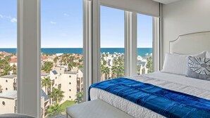 A bedroom with a large window, a bed with a blue bedspread, and a view of a coastal town. - Where the Sky Meets the Sea – Luxury Dream Views (South Padre Island)