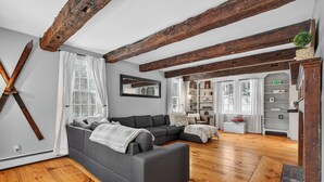 Ein modernes Wohnzimmer mit einem grauen Ecksofa, Holzboden, sichtbaren Holzbalken, einem großen Spiegel und einem Bücherregal.