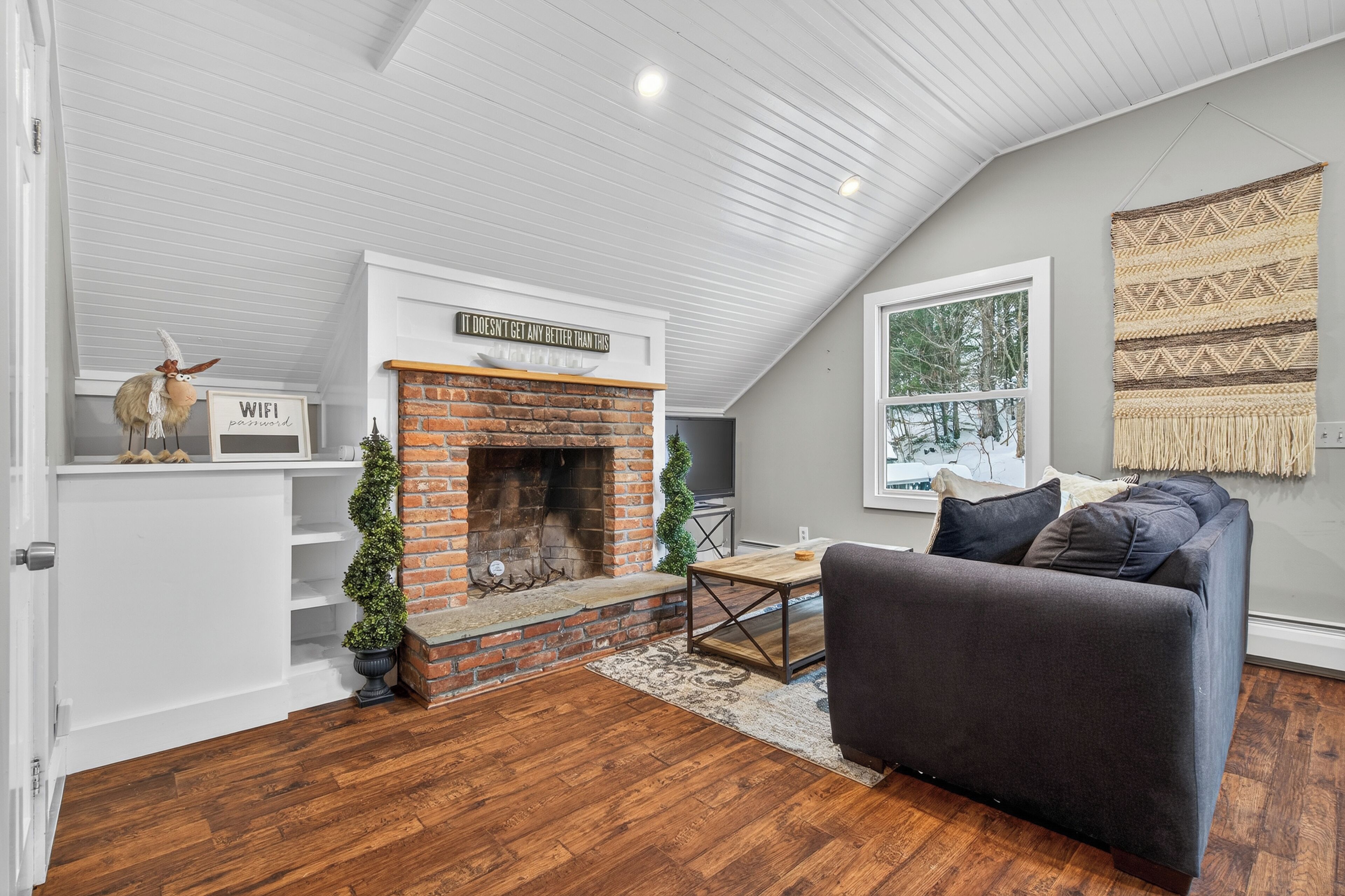 A cozy living room with a brick fireplace, a gray couch, and a large window offering a view of trees.