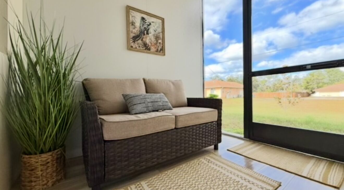 A room with a wicker sofa, a potted plant, a framed artwork, and a view of a green landscape through a large window.