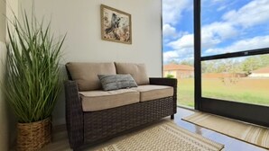 A room with a wicker sofa, a potted plant, a framed artwork, and a view of a green landscape through a large window. - Private Cozy and relaxing 1-bedroom unit with living room kitchen full bath more (North Port)