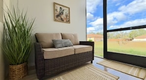 A room with a wicker sofa, a potted plant, a framed artwork, and a view of a green landscape through a large window. - Private Cozy and relaxing 1-bedroom unit with living room kitchen full bath more (North Port)