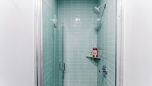 A bathroom with teal tiles, a glass shower enclosure, a handheld showerhead, a glass door, and a shelf with toiletries. - S. Philly Studio Minutes - Center City Passyunk Sq and More! (Philadelphia)