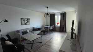 Una sala de estar moderna con un sofá, una mesa de centro y sillas. Hay un comedor con una mesa y sillas. Se ve un ventanal con cortinas.