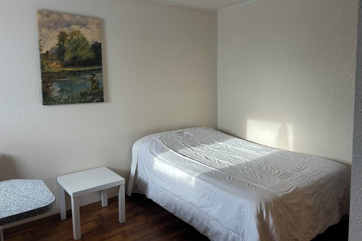 A small, single-bed room with a painting of a landscape on the wall, a small white table, and a wooden floor.