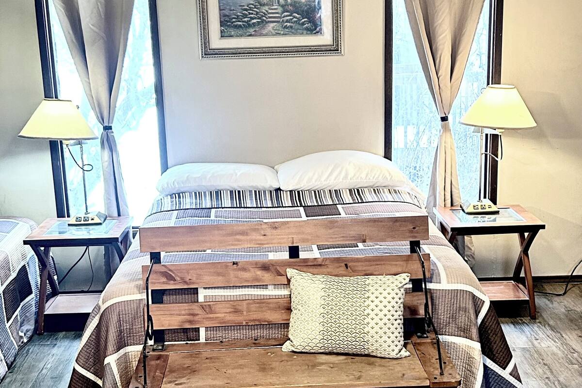 A bedroom with a wooden bed, two bedside tables with lamps, a framed picture on the wall, and a window with curtains.