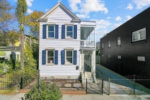 Une maison à deux étages avec un balcon, une cour clôturée et un bâtiment noir situé à côté.
