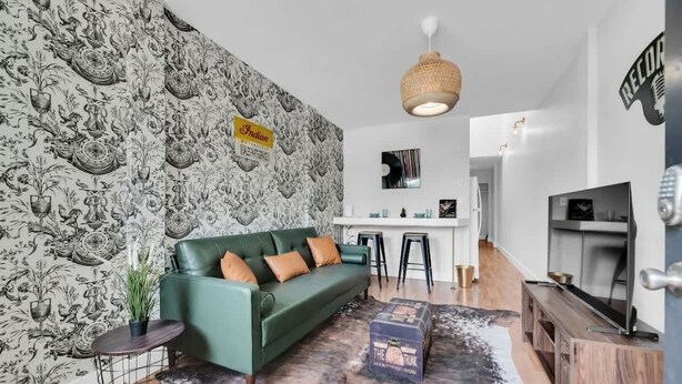 A living room with a green sofa, a small side table with a plant, a patterned wallpaper, a hanging light fixture, and a television on a wooden stand. - NEW WeHo Hotspot | 8 min Broadway, DTN + Music Row (Nashville)