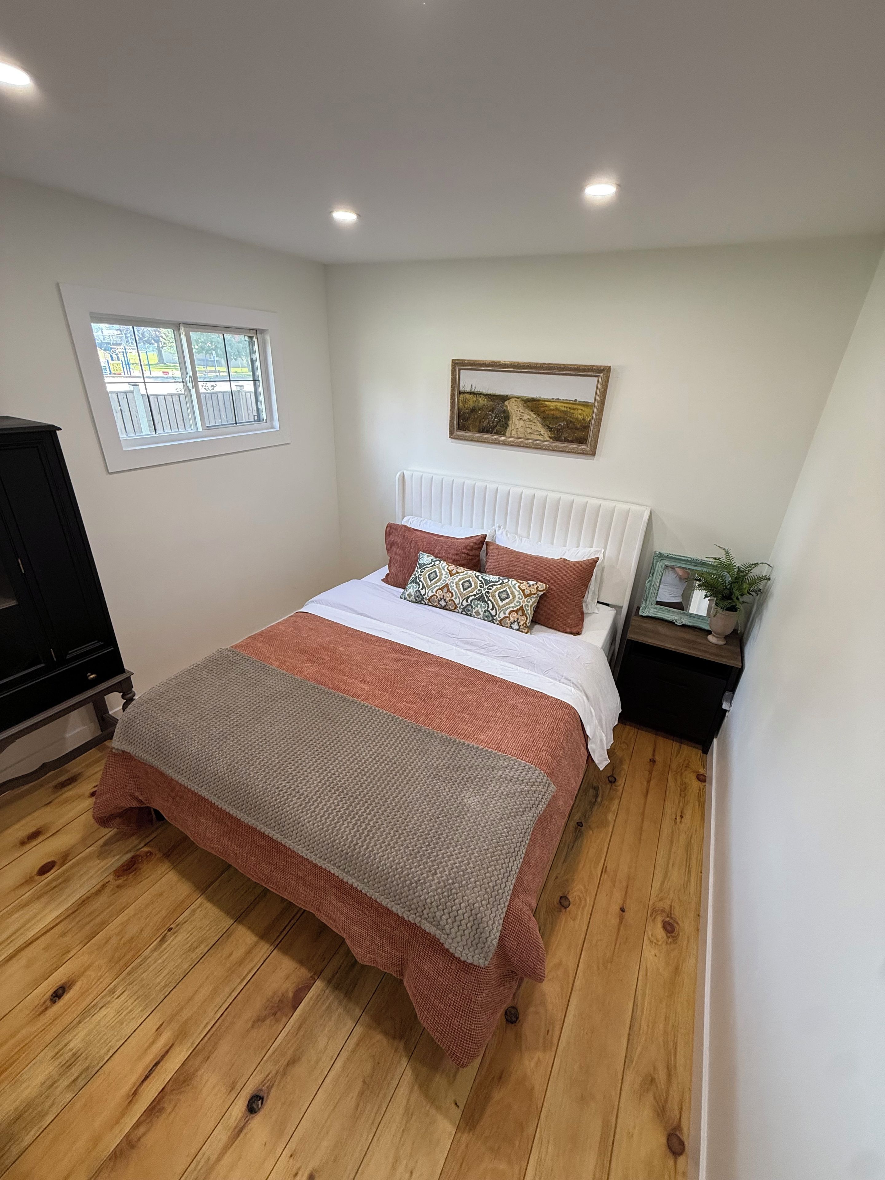 A bedroom with a bed, a window, a radiator, a nightstand, and a plant.