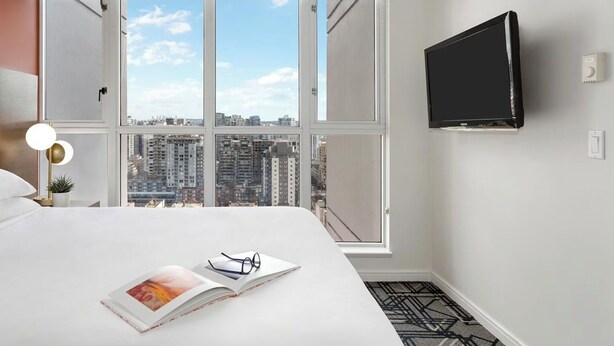 Une chambre d’hôtel moderne dotée d’un grand lit, d’un téléviseur fixé au mur et offrant une vue sur le paysage urbain grâce à de grandes fenêtres.