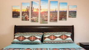 Een bed met gestreepte kussens, een blauwgroene deken en een muur met vijf ingelijste schilderijen van woestijnlandschappen.