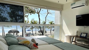 A bedroom with a large window overlooking a beach, a bed with a colorful pillow, and a TV on the wall. - Playa Caracol, a Happy Beach (Punta Chame)
