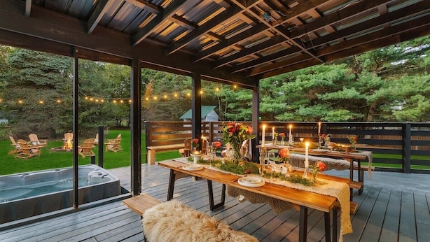 An outdoor deck with wooden tables set for dining, surrounded by chairs, and a hot tub. - Near Ithaca! Hot Tub! Karaoke Room! Sleeps 10! Dogs ok! (Newfield)