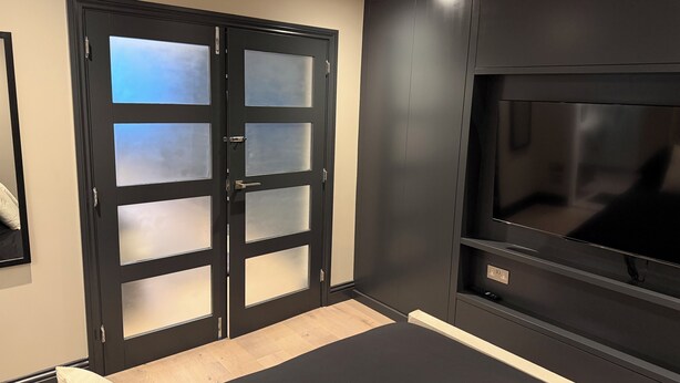 Una habitación con una puerta enmarcada en negro y paneles de vidrio esmerilado, un mueble negro para entretenimiento con un televisor de pantalla plana, y un suelo de madera claro.