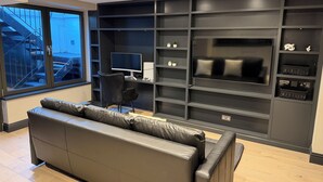 Un salón moderno con un sofá de cuero negro, un televisor de pantalla plana montado en una pared oscura, y un escritorio con un monitor y un teclado.