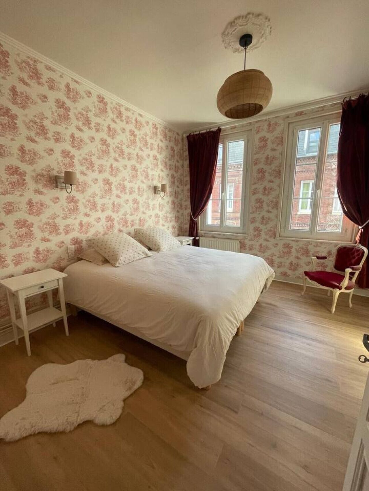 Ein Schlafzimmer mit Bett, Nachttisch, Stuhl, einem Fenster mit Vorhängen und einer Heizung.