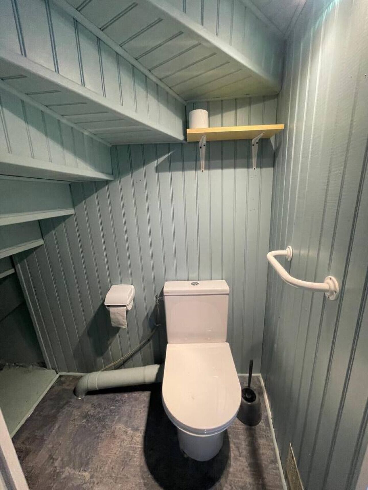 A small, narrow bathroom with a toilet, a handrail, and a shelf above the toilet.