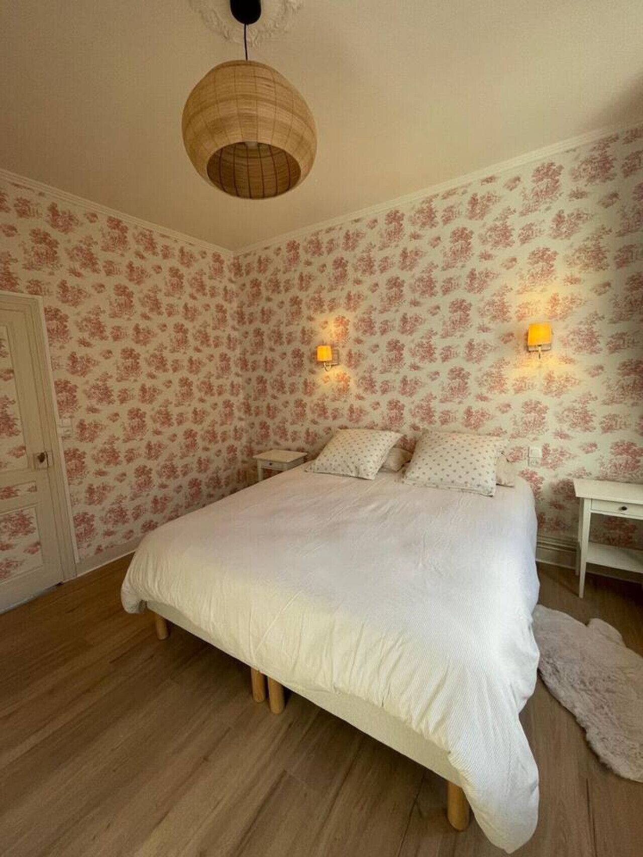 Un dormitorio con una cama grande, papel tapiz floral, dos lámparas de pared y suelo de madera.