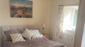 A bedroom with a bed, a nightstand, a lamp, a painting, and a window with curtains. - Swallow Bach, Couples Bolt Hole (Llangefni)