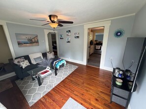 Un salon avec un canapé, une table basse et un ventilateur de plafond. On peut apercevoir une cuisine à travers une porte ouverte.