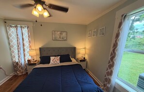 Un dormitorio con cama, dos lámparas de noche, ventilador de techo, ventana con cortinas y una pared con tres cuadros enmarcados.