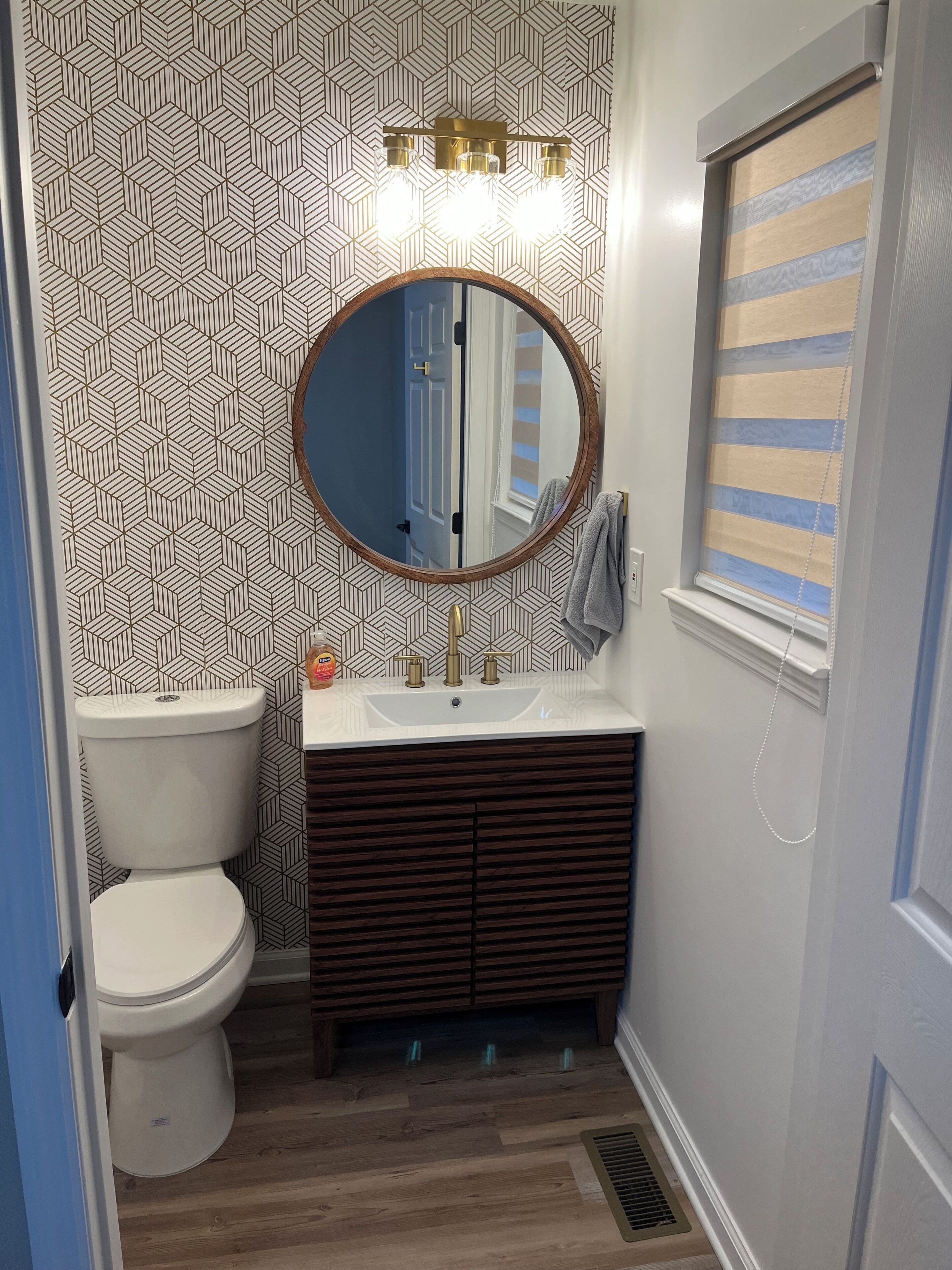 Een badkamer met een wand met een geometrisch patroon, een ronde spiegel, een wit toilet, een houten wastafelmeubel en een raam met een gestreept zonnescherm.