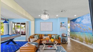 Ein Wohnzimmer mit Billardtisch, braunem Ledersofa, einer Kristallleuchtre und einem Wandbild mit Strandmotiv.