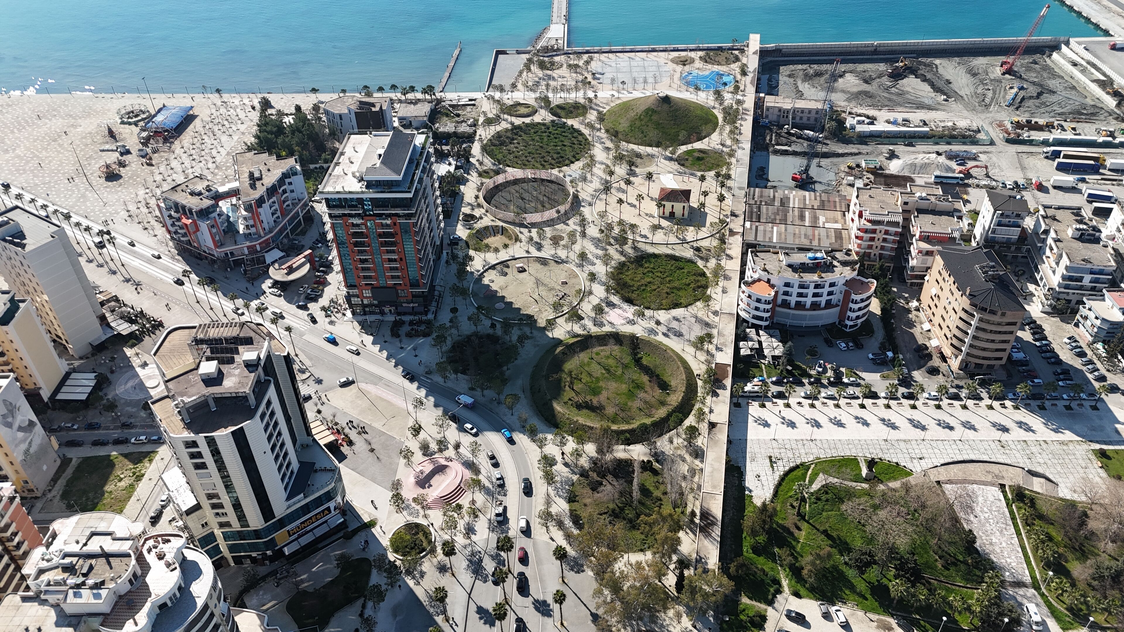 An aerial view of a coastal urban area with modern buildings, a central plaza with circular green spaces, and a beach with umbrellas.