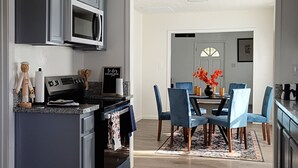 Een moderne keuken met grijze kastjes, een roestvrijstalen magnetron en een eethoek met blauwe stoelen en een vloerkleed met bloemen.