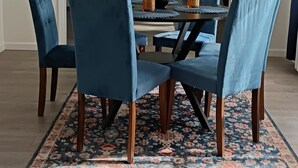 Een eetgedeelte met een ronde tafel, zes blauwe gestoffeerde stoelen, een vloerkleed met bloemenprint en een vaas met oranje bloemen.