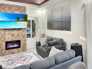 A living room with a stone fireplace, a grey sofa, a flat-screen TV, and a large window with curtains. - Cielo y Mar (Gibson bight)
