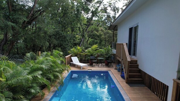 Una piscina con cubierta de madera, rodeada de vegetación exuberante y un edificio blanco con una escalera.