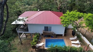 Una casa de dos pisos con techo rojo, piscina y terraza con sillas de descanso.