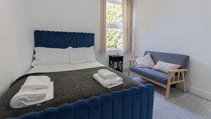 A neatly made bed with white linens, a blue headboard, and a grey blanket. There is a grey sofa with cushions and a small wooden table beside it.